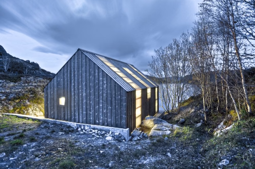 vista trasera Casita de madera oscura en el fiordo al anochecer con luz cálida saliendo por sus ventanas