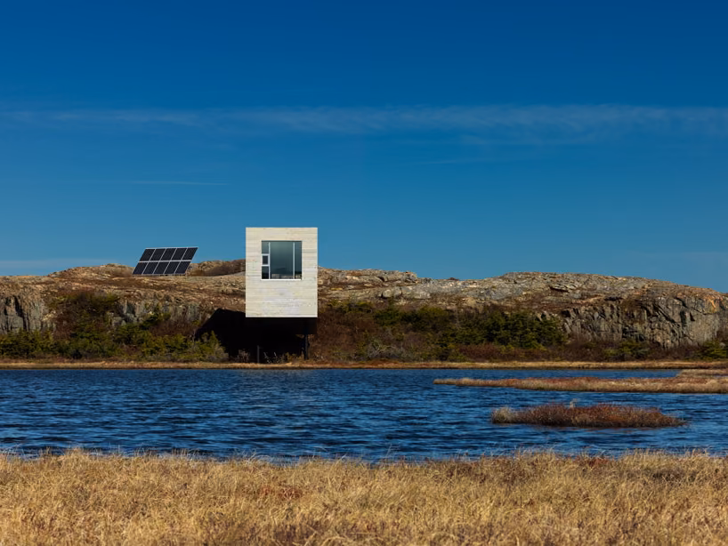 vista de frente Fachada frontal del Bridge Studio de Todd Saunders junto a un lago azul, con paneles solares en la roca