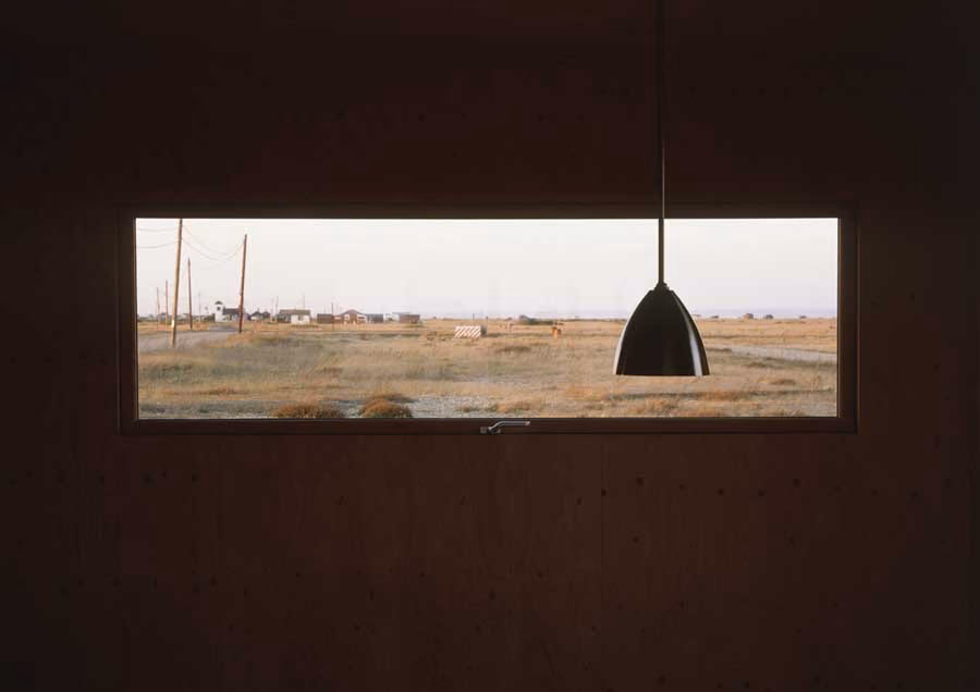 ventana Ventana horizontal en una pared de madera oscura que enmarca el llano de Dungeness, con lámpara colgante