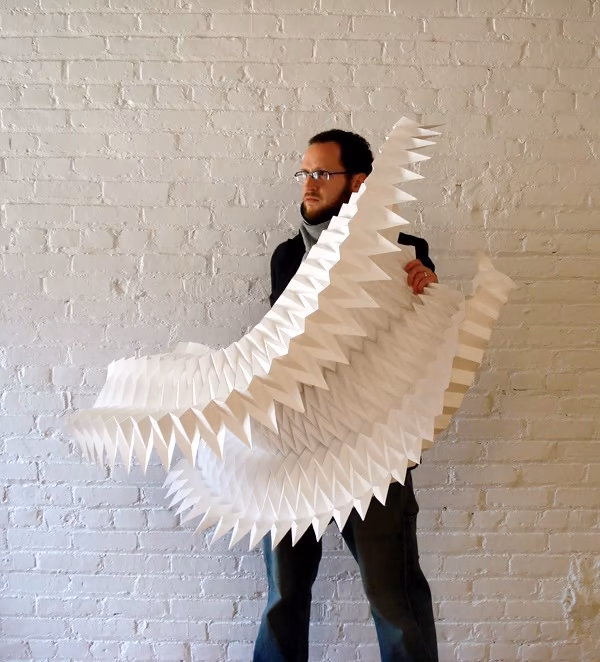 Matthew Shlian Persona sosteniendo una gran escultura de papel blanco plegado en pico de Matthew Shlian ante una pared de ladrillo