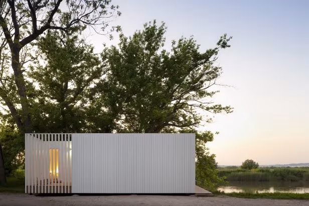 Pabellón blanco de lamas verticales junto a un humedal entre árboles al atardecer en Treehouse Riga