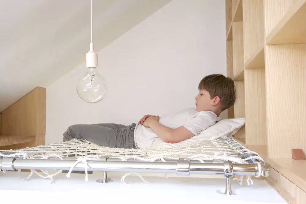 Niño tumbado en una cama de red bajo una bombilla colgante en una habitación de estudiantes con muebles de madera clara