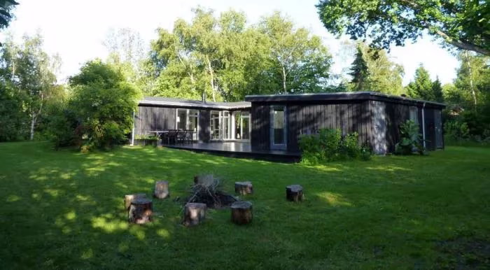 Casa baja de madera oscura al fondo de un jardín danés de césped con tocones y árboles frondosos