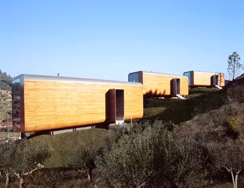 Prototipos transportables de ÁBATON: módulos alargados revestidos de madera sobre una ladera con arbustos y cielo azul