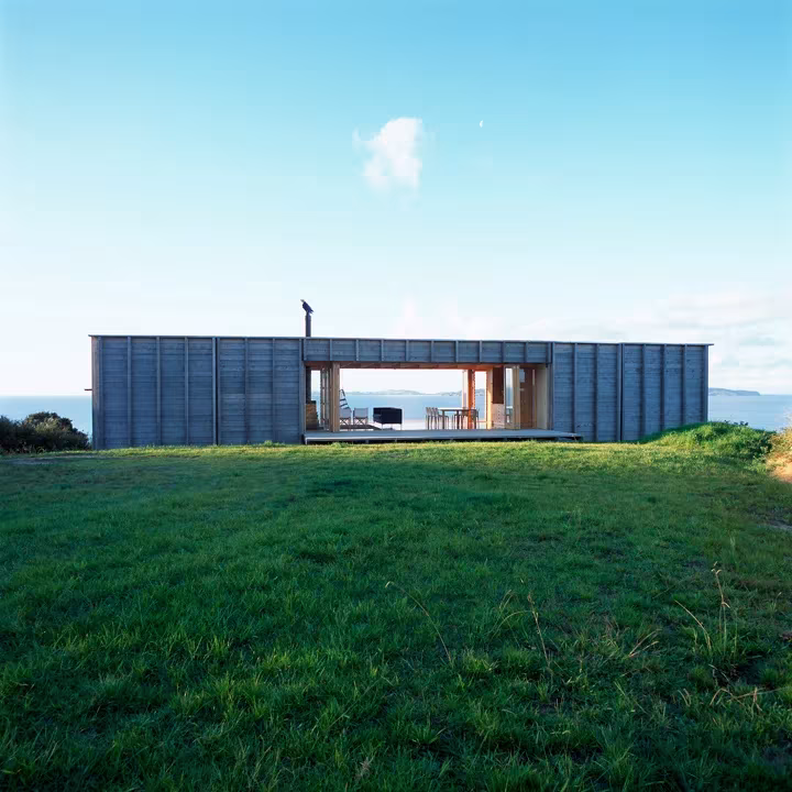Cabaña en Coromandel de Crosson Clarke Carnachan: caja de madera abierta en el centro sobre un prado verde frente al mar