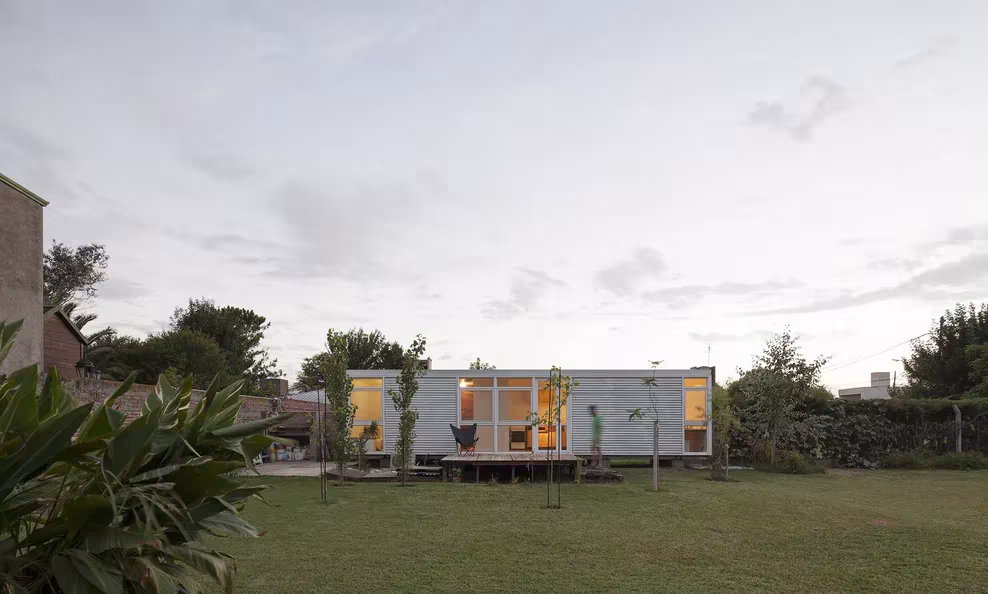Mia House en Argentina: pequeño volumen de chapa con ventanas iluminadas al fondo de un jardín de césped