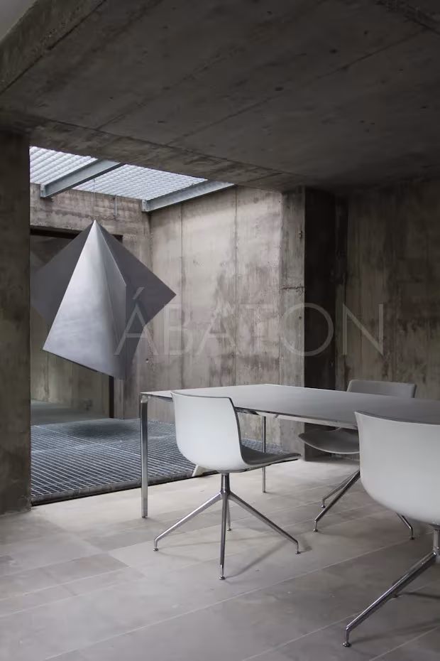 Oficina-1 Comedor de hormigón visto con mesa blanca y escultura plegada de metal bajo un lucernario, en la oficina-vivienda