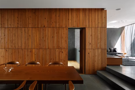 Salón con muro revestido de listones de madera y mesa de comedor ante una puerta corredera, en la Optical Glass House