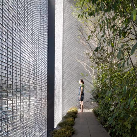 Muro de bloques de cristal de la Optical Glass House junto a un sendero con arbustos y una persona mirando hacia arriba