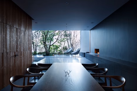 Comedor de la Optical Glass House con mesa larga y paredes de madera oscura abierto al jardín