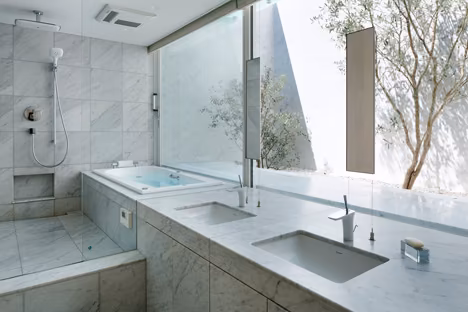 Baño de mármol claro con bañera y lavabos dobles ante un ventanal que da a un patio con árboles