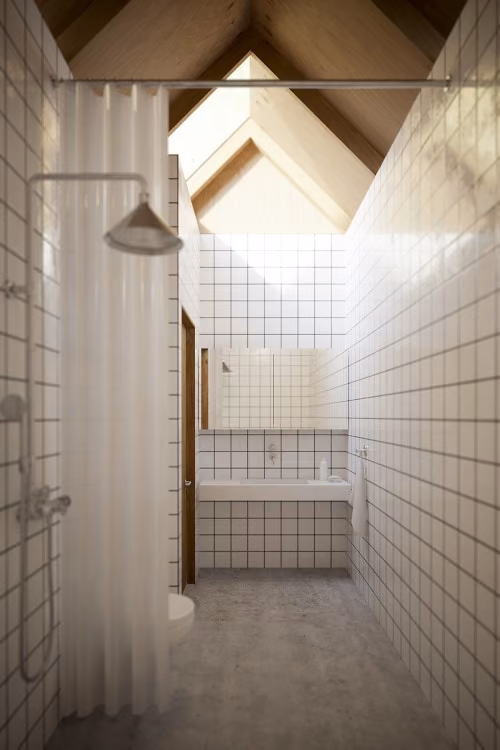 Baño de azulejo blanco bajo la cubierta inclinada de madera y su lucernario en la Casa para Madre