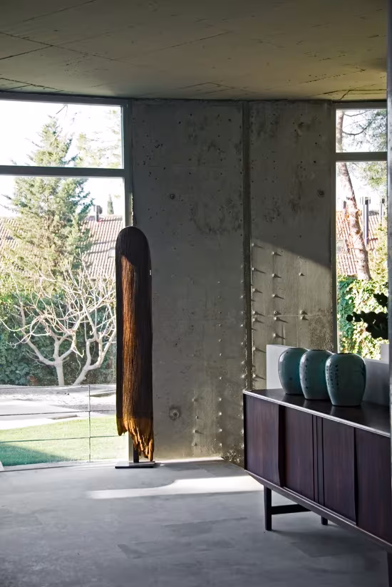 Esquina del salón con pilar de hormigón visto, aparador oscuro y cristalera sobre el césped del jardín