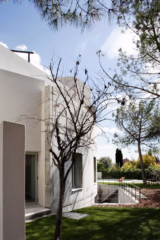Fachada blanca de la vivienda semicircular entre pinos, con escalera hacia el patio inferior ajardinado