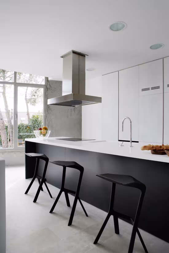 Cocina blanca con isla negra, taburetes y campana de acero inoxidable ante ventanas arboladas