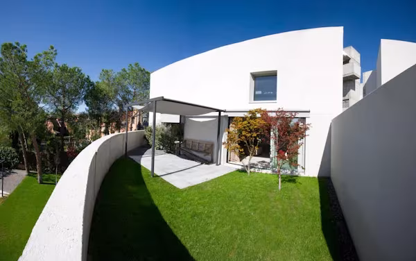Jardín en cubierta con muro curvo de hormigón y pérgola ligera ante una vivienda blanca rodeada de pinos