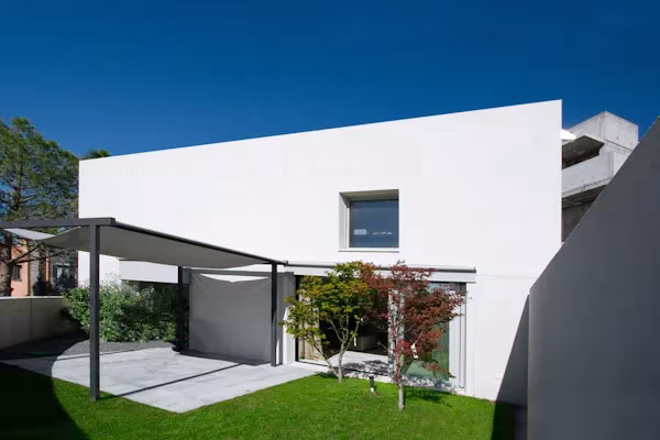 Terraza con césped bajo una pérgola de toldo ante un volumen blanco con ventana pequeña