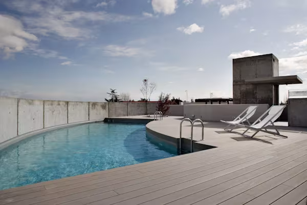 Piscina de borde curvo con tarima gris y tumbonas sobre una azotea, con la torre de hormigón y el cielo al fondo