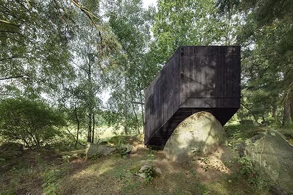 Cabaña en Bohemia de Uhlik Architekti: volumen de madera negra apoyado sobre una roca en un claro del bosque