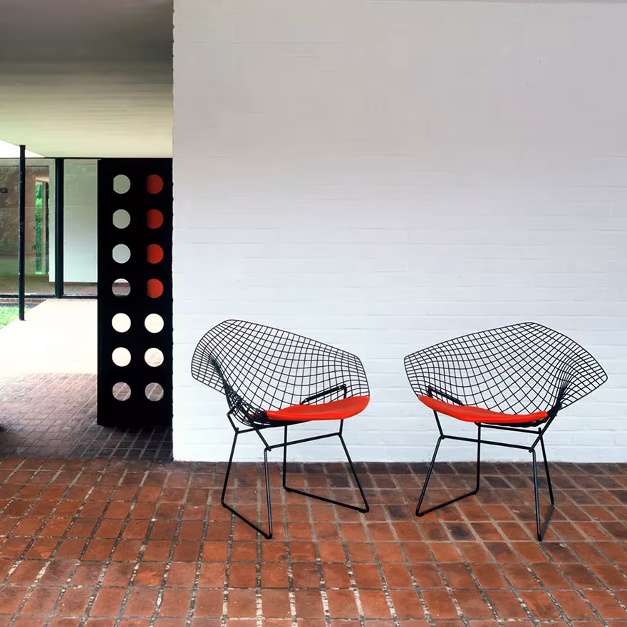 Dos sillas Diamond de Harry Bertoia con cojín rojo sobre un suelo de ladrillo junto a una pared blanca