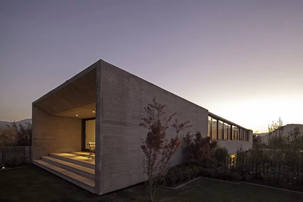 Casa SH de 01ARQ, volumen de hormigón con un porche abierto iluminado sobre el jardín al atardecer