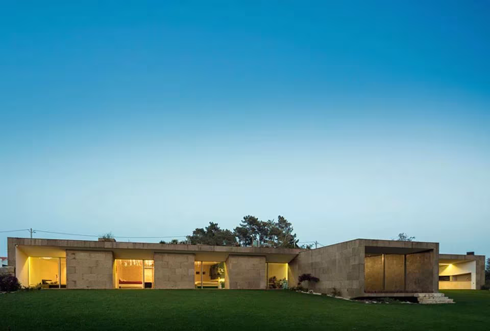 Hill Cork House de Contaminar Arquitectos, volumen bajo revestido de corcho con ventanas iluminadas al anochecer