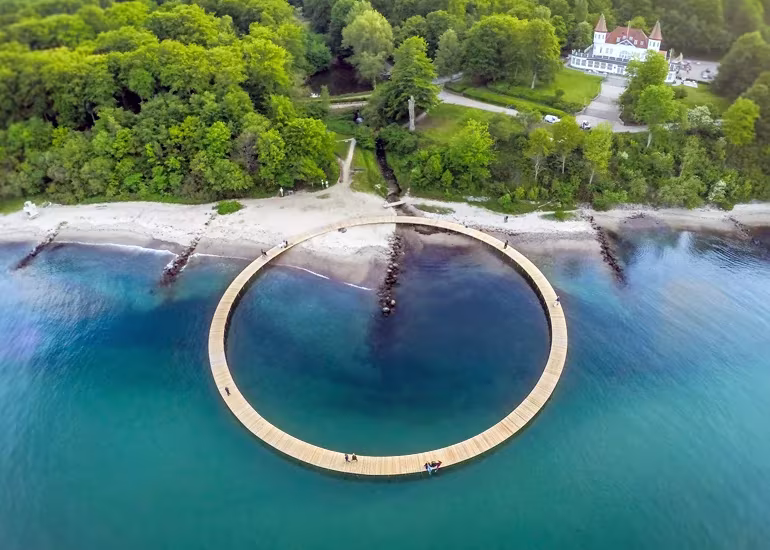Vista aérea del puente circular de madera de Aarhus sobre el mar turquesa, junto a una playa y un bosque