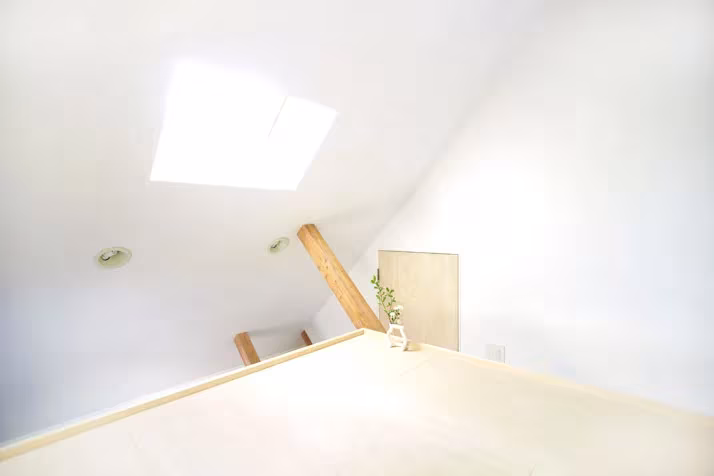 Altillo blanco bajo la cubierta inclinada de la Casa H, con lucernario, puerta pequeña y una viga de madera