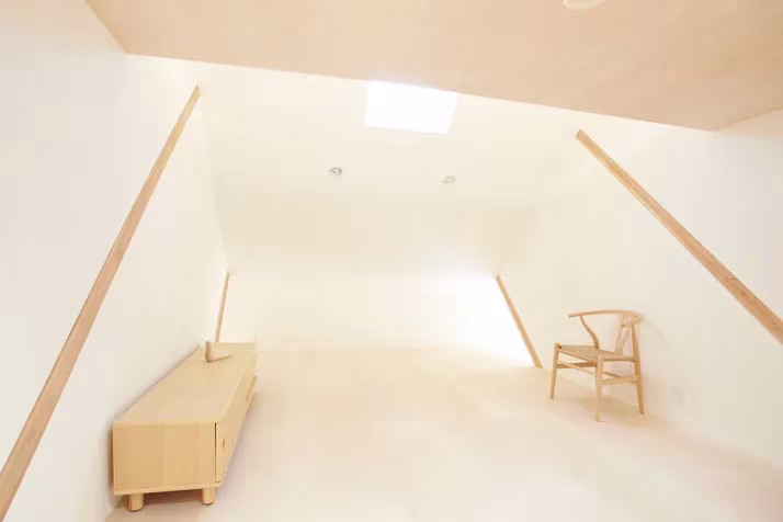 Estancia blanca de la Casa H con techo inclinado, lucernario, un mueble bajo de madera y una silla
