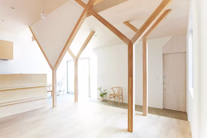 Interior blanco de la Casa H con columnas de madera en forma de Y, puerta de madera y una silla junto a la pared