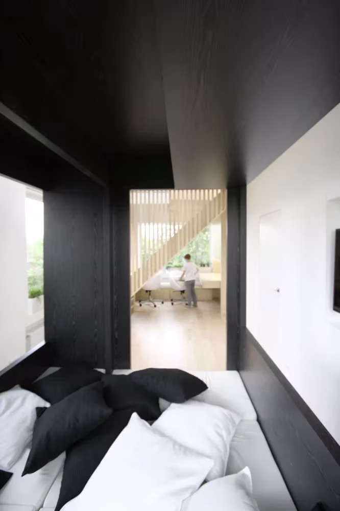 Dormitorio dentro del cubo negro transformable, con cojines blancos y negros y vista al cuarto