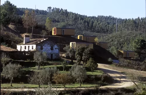 Módulos prefabricados transportables junto a casas de piedra en una ladera rural con olivos y bosque