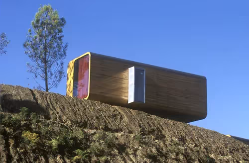 Módulo prefabricado transportable de madera con frente de cristal posado sobre un terreno arado junto a un pino