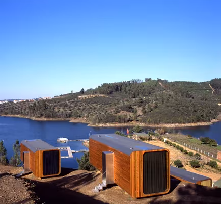 Dos módulos prefabricados de madera en una ladera frente a un embalse rodeado de colinas con olivos