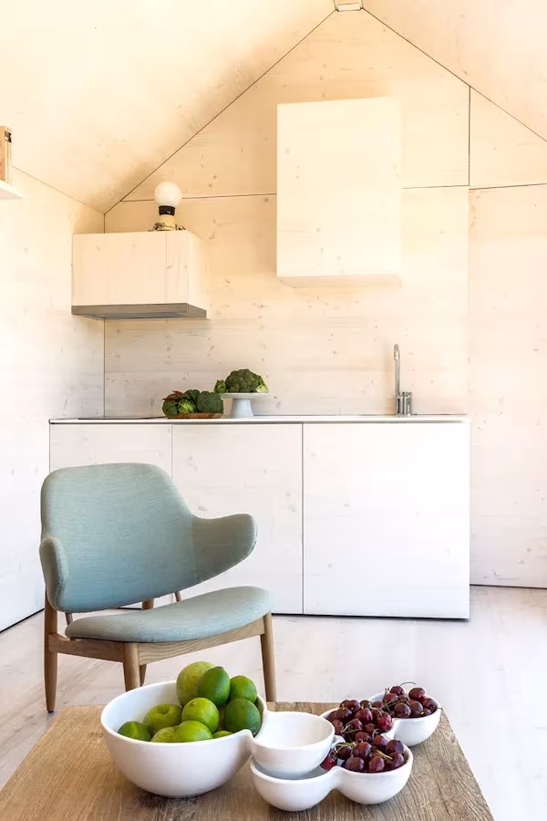 Cocina blanca de la casa ÁPH80 de ÁBATON bajo techo a dos aguas, con butaca azul y cuencos de fruta