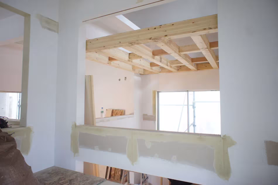 Interior de la House T en construcción con paredes blancas, entramado de madera en el techo y ventanas