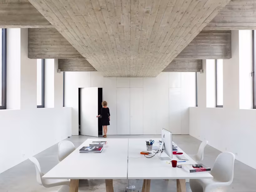 Sala blanca bajo el forjado de hormigón de la piscina de dmvA en Flandes, con mesa de trabajo y sillas