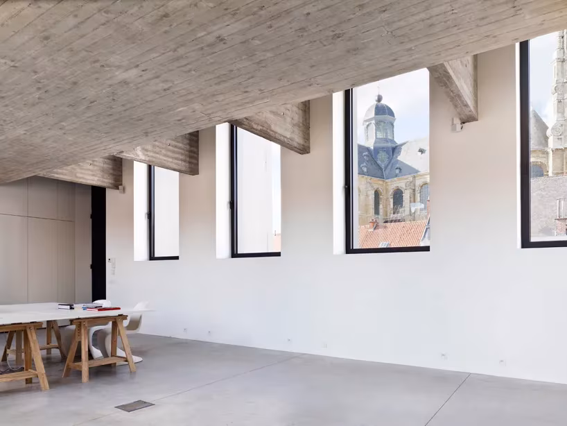 Estancia diáfana de suelo gris con ventanas verticales que enmarcan una torre de iglesia flamenca