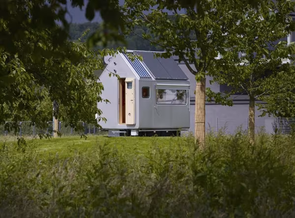 Cabina Diogene de Renzo Piano entre árboles y hierba alta, con ventanas laterales y paneles grises