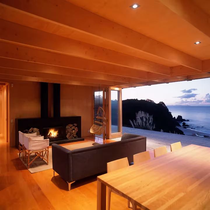 Salón de la cabaña de Crosson Clarke Carnachan con chimenea, sofá oscuro y pared abierta al mar