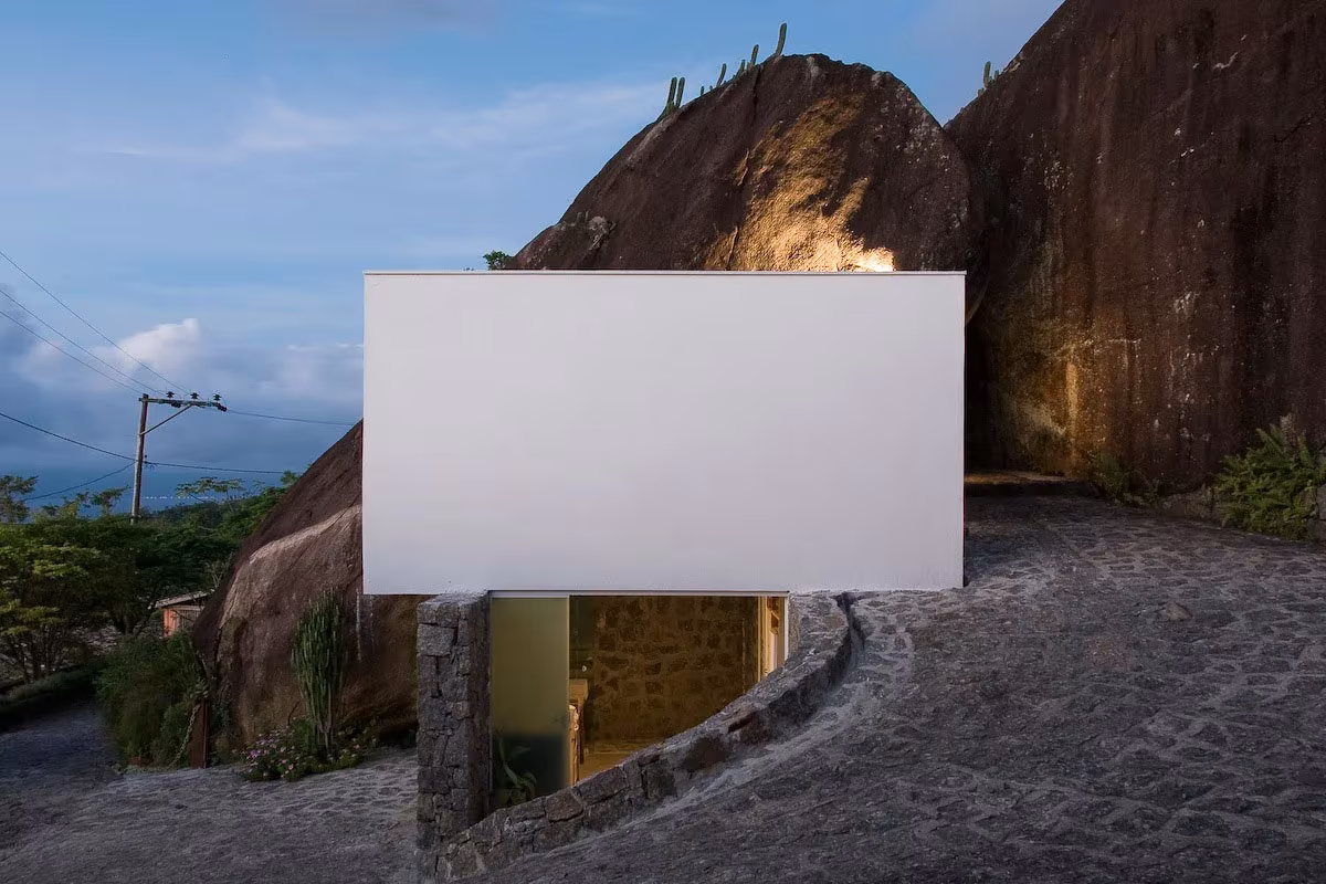 Box House: caja blanca de hormigón suspendida entre dos grandes rocas al anochecer en una isla de Brasil