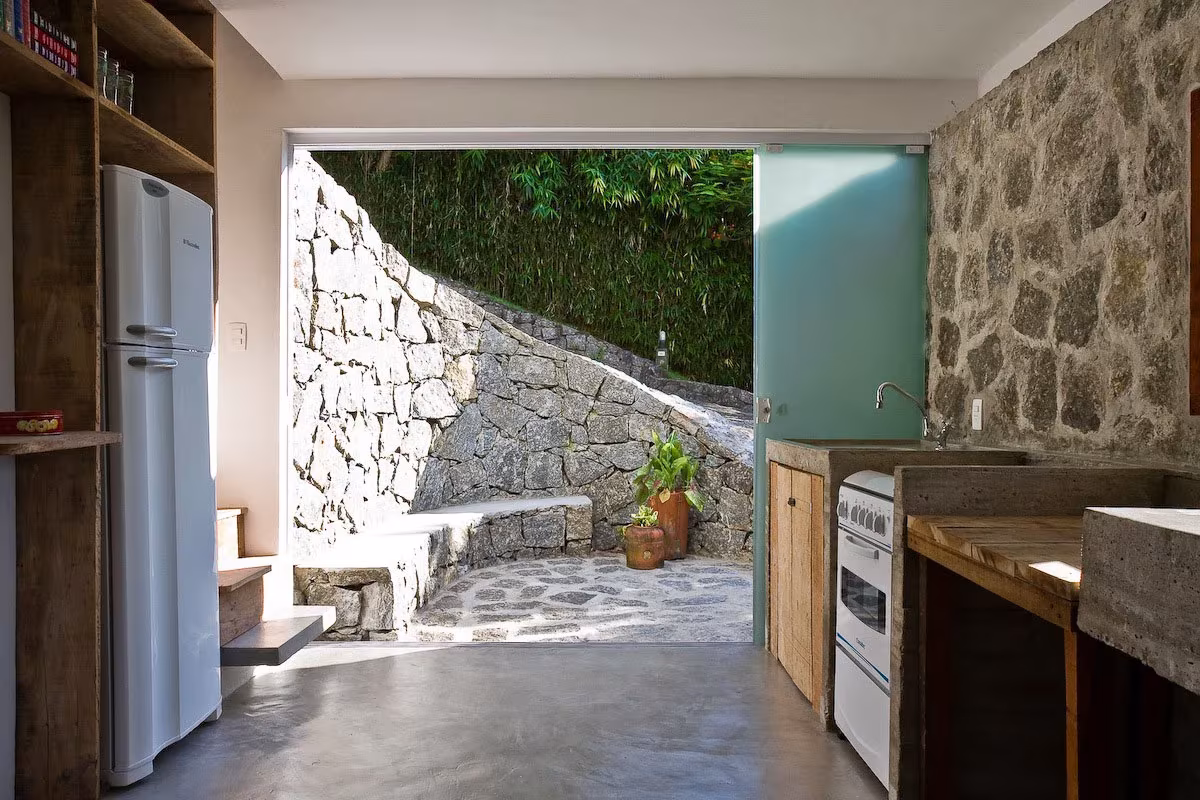 Cocina de la Box House con frigorífico, muros de piedra y puerta abierta a un patio empedrado con bancos