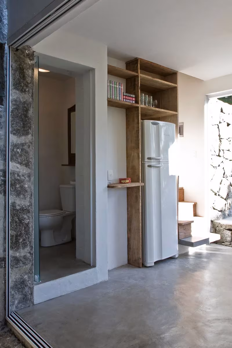 Interior de la Box House con suelo de cemento, baño, frigorífico y estantes de madera junto a muros de piedra