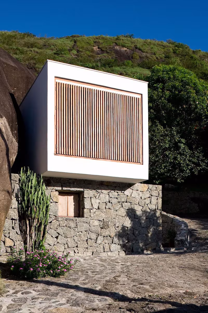Caja blanca de hormigón con celosía de lamas de madera sobre el muro de piedra de la Box House