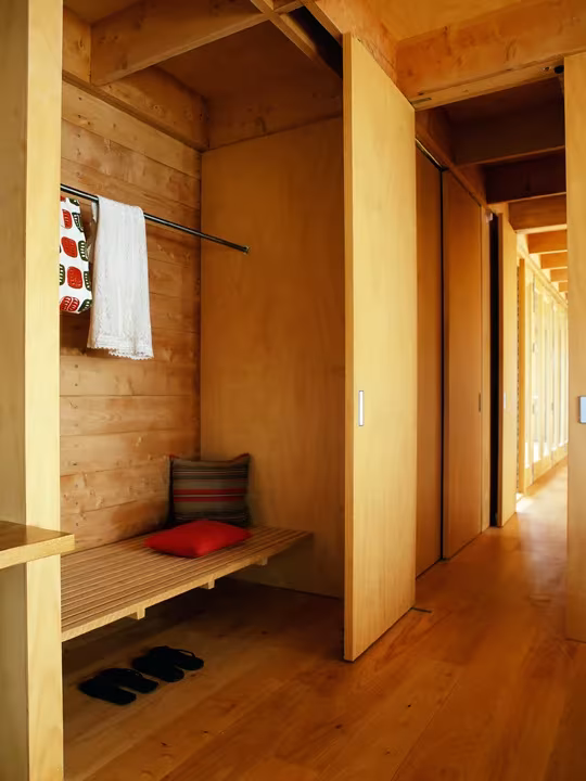 Pasillo y vestidor revestidos de madera en la cabaña de Coromandel, con banco, perchero y luz cálida