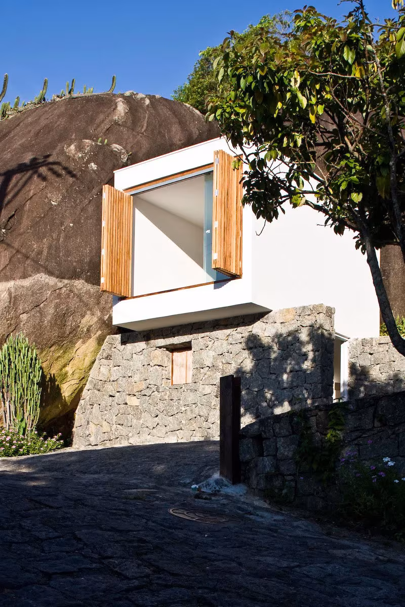 Caja blanca de la Box House con contraventanas de madera abiertas, encajada junto a una gran roca