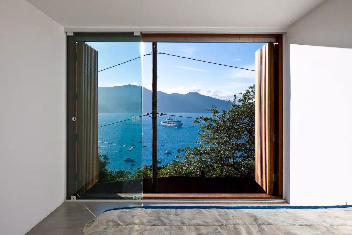 Gran ventana del dormitorio de la Box House abierta a la bahía, con barcos y montañas al fondo