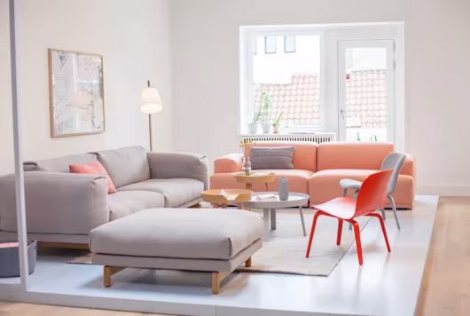 Salón luminoso de Muuto con sofás grises y salmón, puf y sillas rojas sobre una alfombra clara