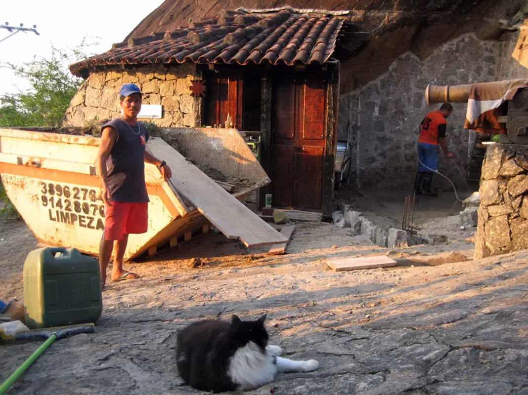 Obras de la Box House: trabajador junto a un contenedor y la casa de piedra original, con un gato tumbado