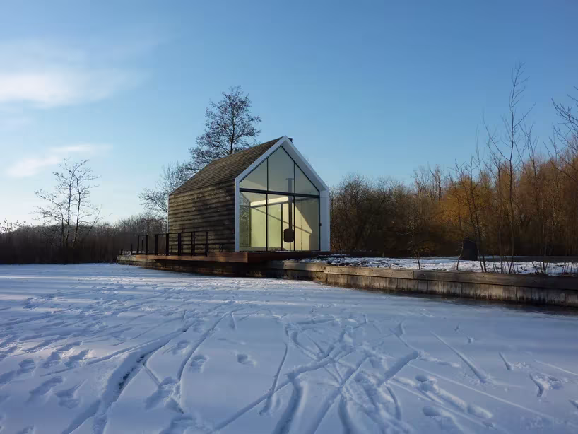 Casita de fin de semana de madera oscura entre árboles desnudos, vista desde un campo nevado